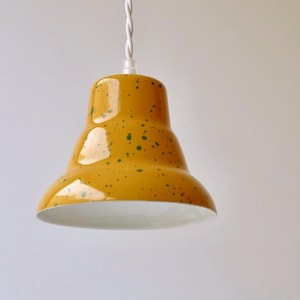 Puede incluir: Una lámpara colgante en forma de campana con un acabado amarillo mostaza brillante y motas de color verde oscuro. El interior de la pantalla es blanco y cuelga de un cable de tela trenzado blanco unido a la parte superior de la pantalla.