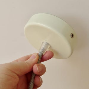 Pode incluir: Rosácea de teto creme com um cabo elétrico trançado cinza. A rosácea é redonda e tem um parafuso na lateral. O cabo é segurado por uma mão.