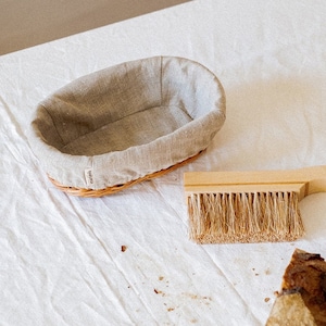 May include: A wooden bread brush with a natural bristle brush head and a leather loop handle. A wicker bread basket lined with a linen cloth and a loaf of bread are also in the image.