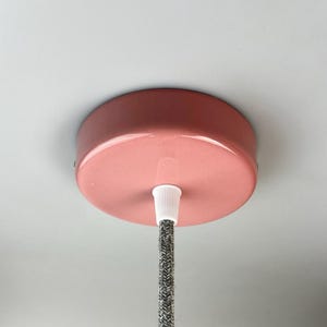 Può includere: Un rosone da soffitto circolare, lucido e di colore rosa polvere per un apparecchio di illuminazione. È dotato di un serracavo centrale in plastica bianca che fissa un cavo elettrico rivestito in tessuto grigio e bianco che scende dal centro.