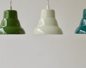 May include: Three glossy pendant lights with tiered, bell-shaped shades in shades of forest green, pale cream, and deep teal. Each lamp features a white cord and a white socket cap, hanging vertically in a row against a plain background.