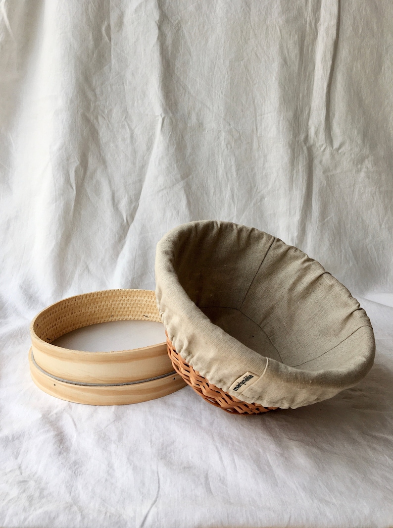 May include: Two round, woven baskets. One is a brown wicker basket with a beige linen liner. The other is a wooden basket with a woven top. The baskets are on a white fabric background.