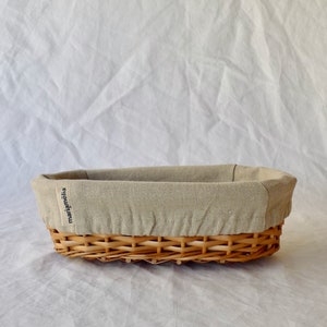 May include: A woven wicker bread basket with a beige linen liner. The basket has a label that reads "mariamelia".