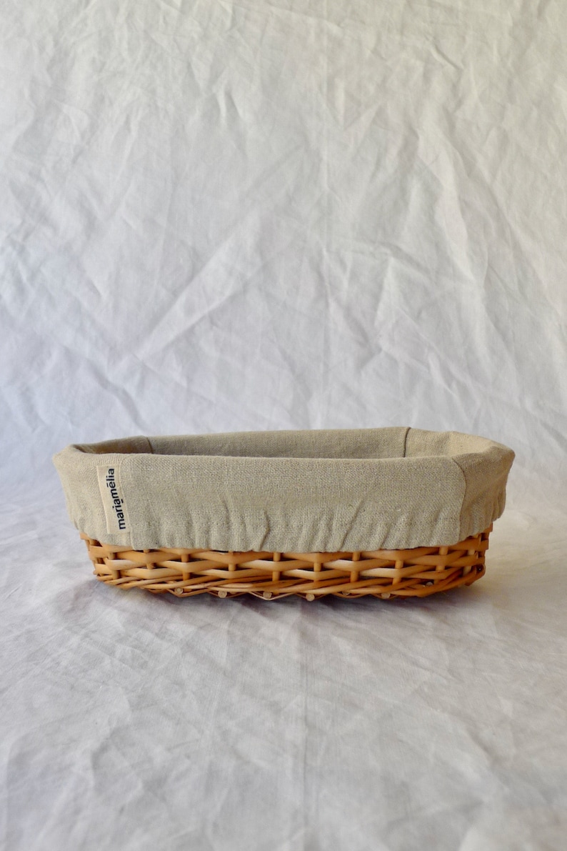 May include: A woven wicker bread basket with a beige linen liner. The basket has a label that reads "mariamelia".