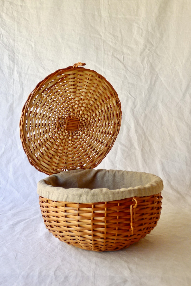 May include: A round wicker basket with a lid, the lid is open and the basket is lined with a light brown fabric.