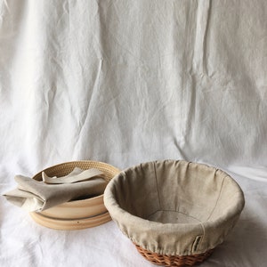 May include: Two round, woven baskets for baking bread. One basket is made of wood and has a linen liner. The other basket is made of wicker and has a linen liner. Both baskets are on a white surface.