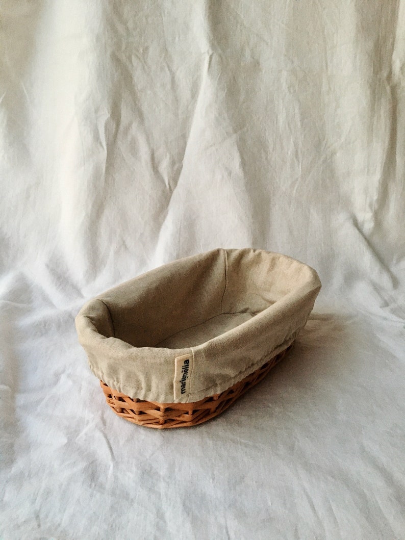 May include: A beige oval-shaped wicker bread basket with a removable beige linen liner. The basket has a label that reads "meijimeila".
