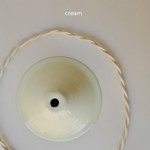 May include: A cream-colored, cone-shaped pendant light canopy with a central opening, accompanied by a matching cream-colored twisted fabric electrical cord arranged in a circular shape. The word cream is printed above the canopy.