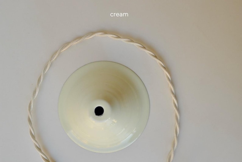 May include: A cream-colored, cone-shaped pendant light canopy with a central opening, accompanied by a matching cream-colored twisted fabric electrical cord arranged in a circular shape. The word cream is printed above the canopy.