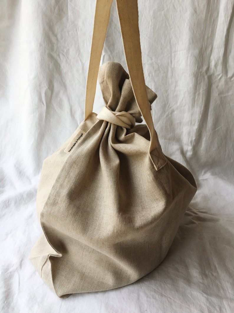 May include: A large, beige linen tote bag with a single long strap. The bag is gathered at the top and tied with a knot. The bag has a small label that says "Handmade in Portugal".