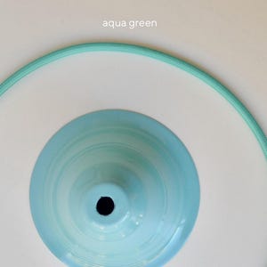 May include: A light blue metal pendant light shade with a circular opening at the center, surrounded by a flexible aqua green fabric-covered electrical cord arranged in a large circle. The text aqua green is printed at the top center.