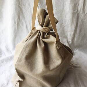 May include: A large, beige linen tote bag with a single long strap. The bag is gathered at the top and tied with a knot. The bag has a small label that says "Handmade in Portugal".