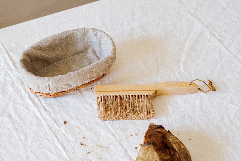 May include: A wooden bread brush with a natural bristle brush head and a leather loop handle. A wicker bread basket lined with a linen cloth and a loaf of bread are also in the image.