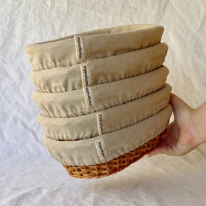 May include: A stack of five beige linen bread baskets with a woven wicker base. Each basket has a black label with the text "mariamélia" printed on it.