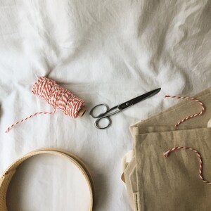 May include: A flat lay of craft supplies including a spool of red and white twine, a pair of silver scissors, a wooden embroidery hoop, and three beige linen bags.