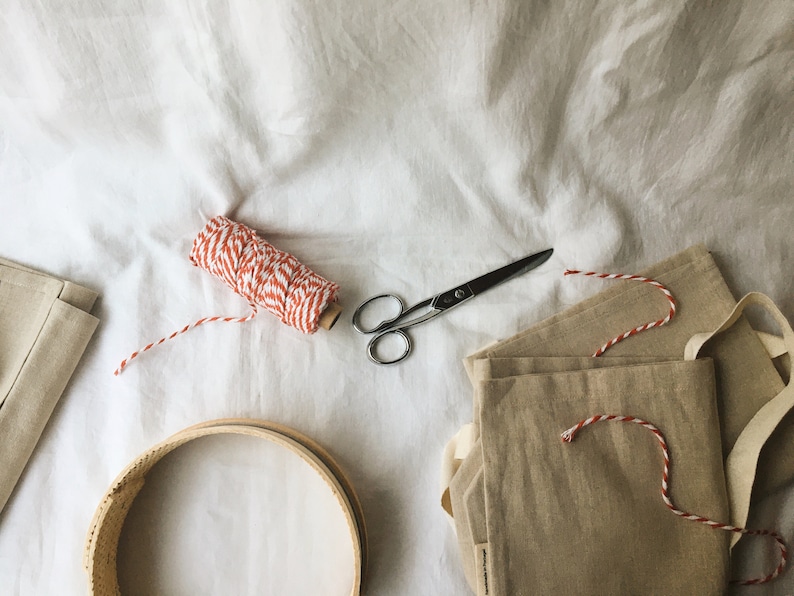 May include: A flat lay of craft supplies including a spool of red and white twine, a pair of silver scissors, a wooden embroidery hoop, and three beige linen bags.