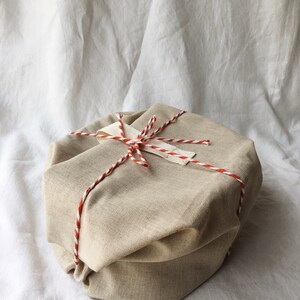 May include: A beige fabric gift box tied with a red and white twine. The box is sitting on a white fabric background.