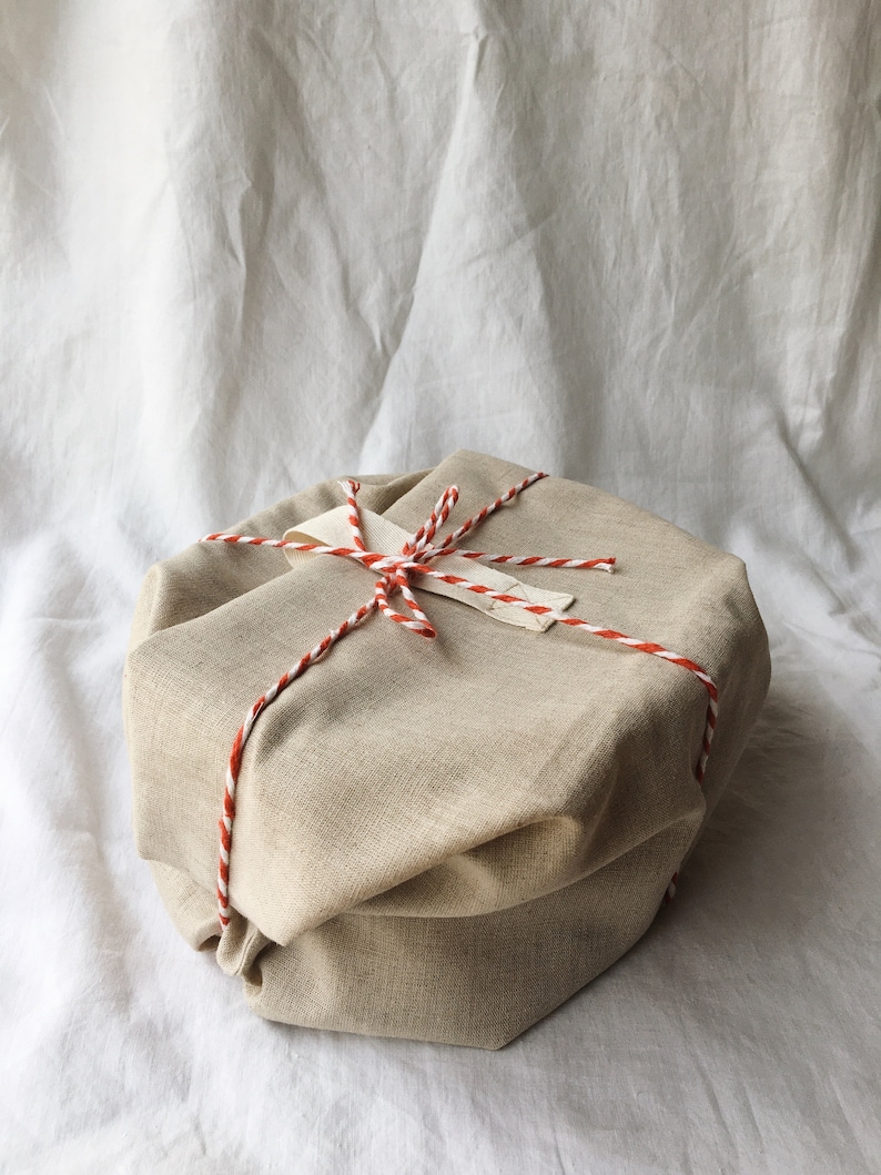 May include: A beige fabric gift box tied with a red and white twine. The box is sitting on a white fabric background.