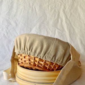 May include: A beige linen cloth cover for a round wooden steamer basket and a woven wicker basket. The cloth cover has a drawstring closure.