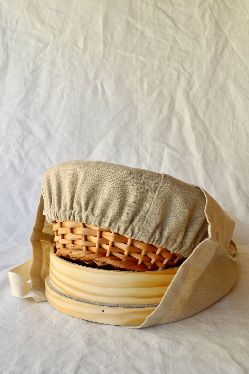 May include: A beige linen cloth cover for a round wooden steamer basket and a woven wicker basket. The cloth cover has a drawstring closure.