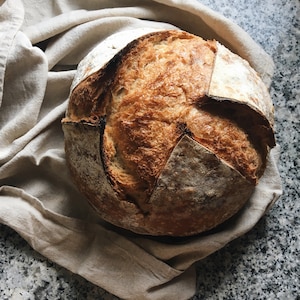 Pode incluir: Uma hoga&ccedil;a de p&atilde;o de fermenta&ccedil;&atilde;o natural rec&eacute;m-assada com uma crosta crocante e um interior macio e arejado. O p&atilde;o est&aacute; descansando sobre um pano de linho bege.