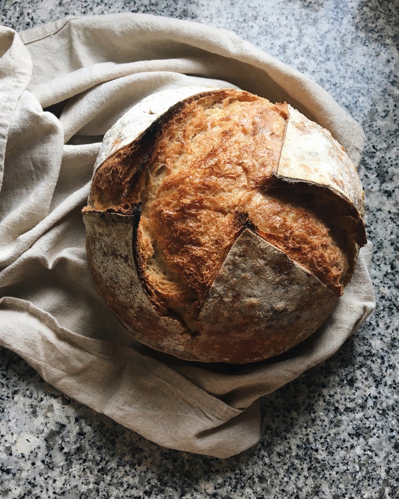 Pode incluir: Uma hoga&ccedil;a de p&atilde;o de fermenta&ccedil;&atilde;o natural rec&eacute;m-assada com uma crosta crocante e um interior macio e arejado. O p&atilde;o est&aacute; descansando sobre um pano de linho bege.