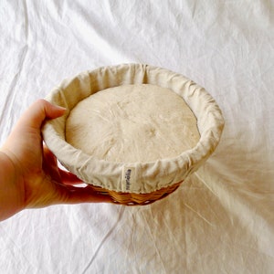 May include: A round, white, linen-lined proofing basket with a light brown dough rising inside. The basket is made of woven wicker and has the word "mj&auml;ill&auml;" printed on the side.