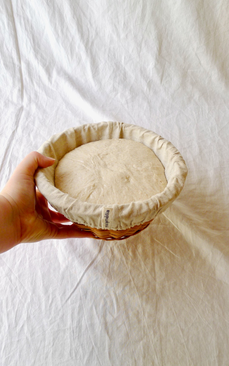 May include: A round, white, linen-lined proofing basket with a light brown dough rising inside. The basket is made of woven wicker and has the word "mj&auml;ill&auml;" printed on the side.