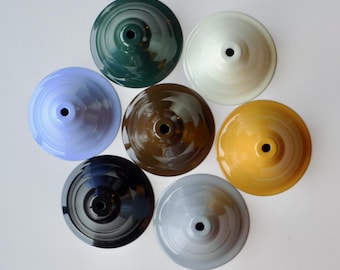 May include: A collection of seven circular, cone-shaped lamp shades arranged in a cluster. The shades feature a smooth, glossy finish in various solid colors including dark green, cream, light blue, brown, black, yellow, and light gray.