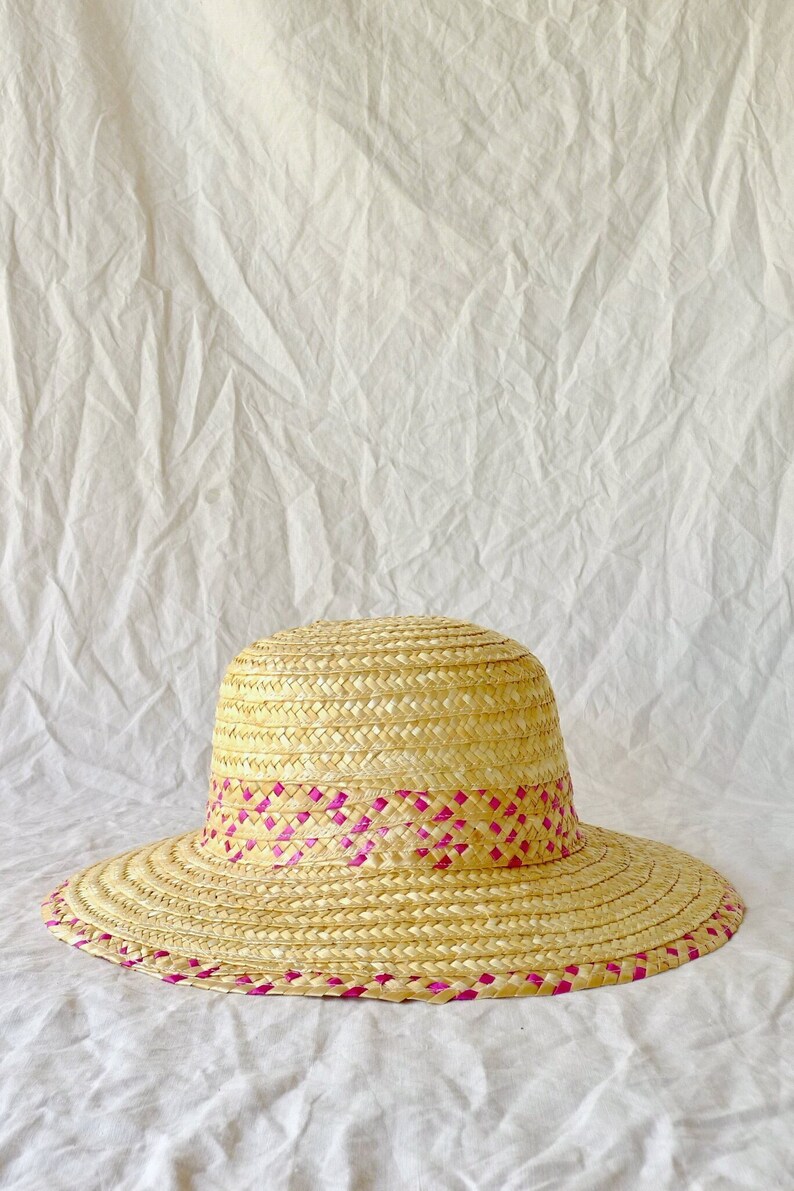 Puede incluir: Un sombrero de paja de ala ancha con una banda tejida rosa alrededor de la corona.