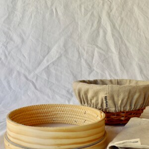 May include: Two round, woven baskets for baking bread. One is a traditional banneton with a linen liner and the other is a wooden proofing basket. Both baskets are on a white linen background.