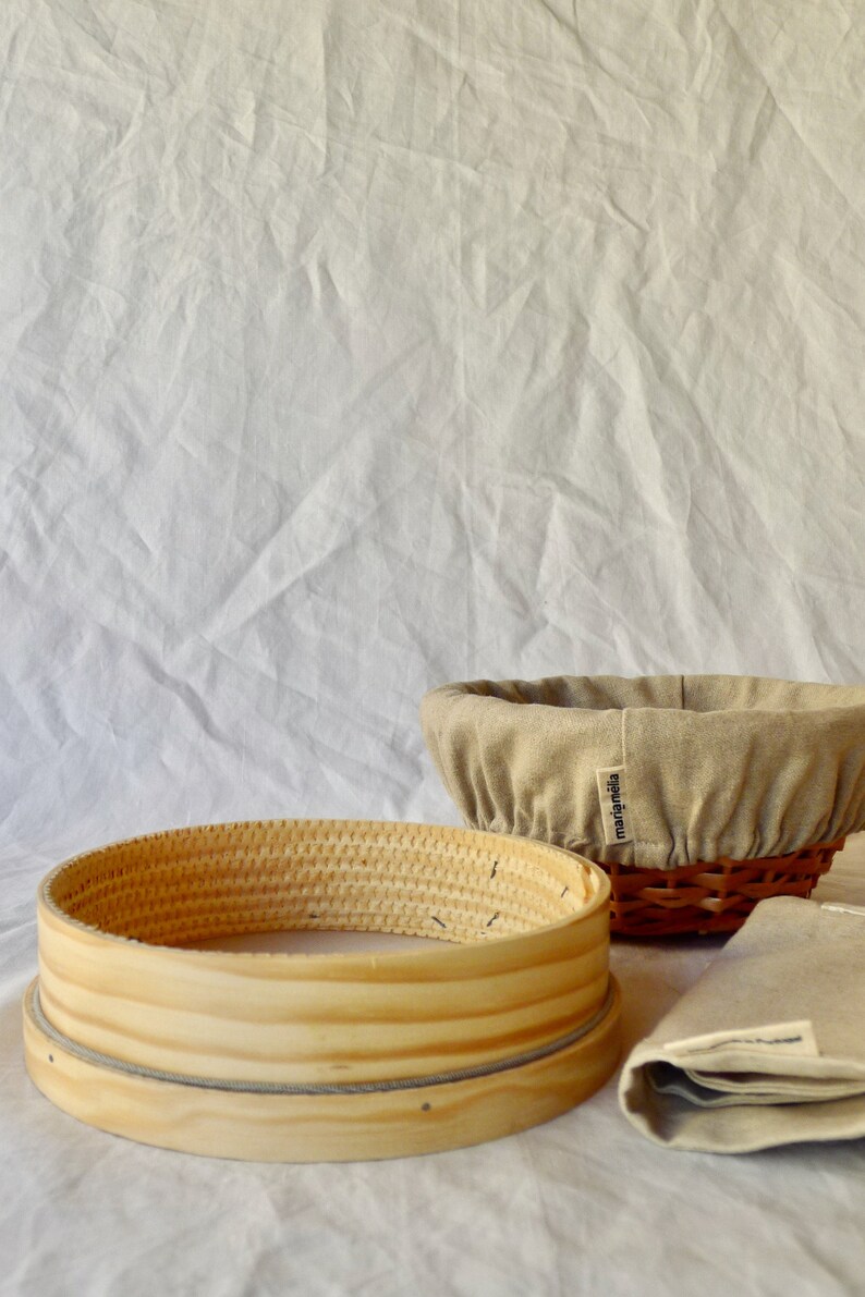 May include: Two round, woven baskets for baking bread. One is a traditional banneton with a linen liner and the other is a wooden proofing basket. Both baskets are on a white linen background.