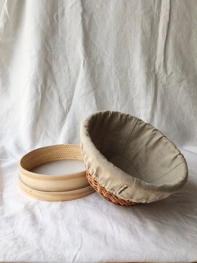 May include: A round, wooden proofing basket with a removable liner. The basket is made of woven wicker and has a light brown color. The liner is made of a light beige fabric and has a drawstring closure.