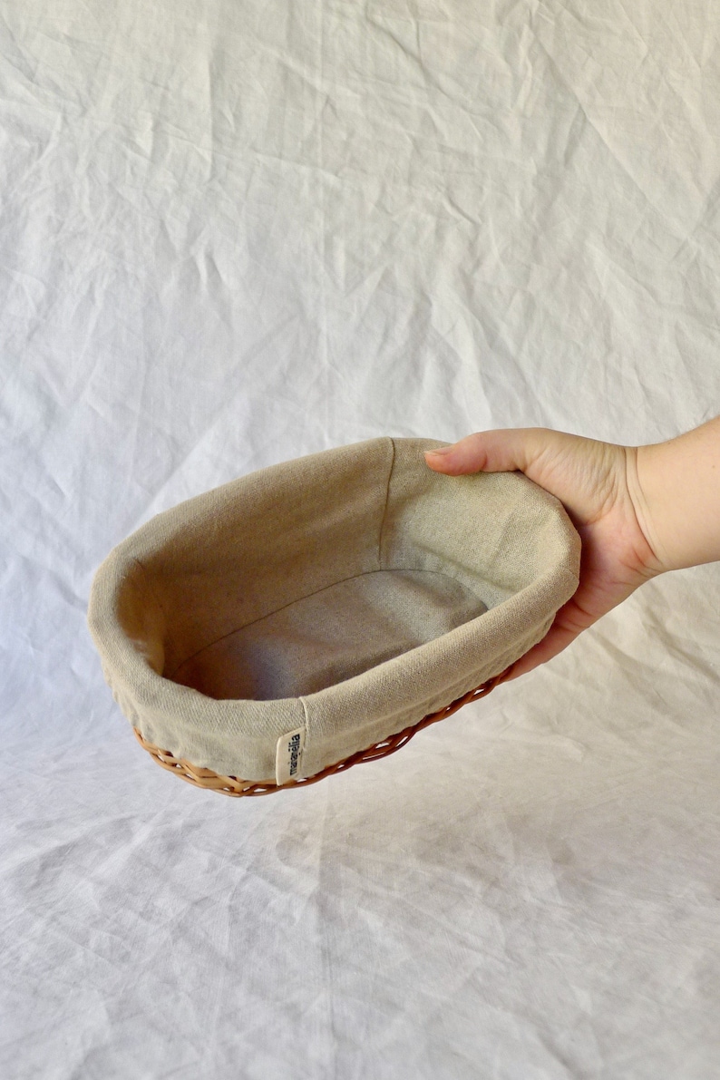 May include: A beige oval-shaped bread basket with a woven wicker base and a removable linen liner. The basket is labeled "marelli."