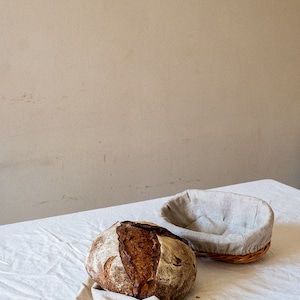 May include: A freshly baked loaf of sourdough bread sits on a white linen tablecloth, next to a brown wicker bread basket lined with a light gray linen cloth.