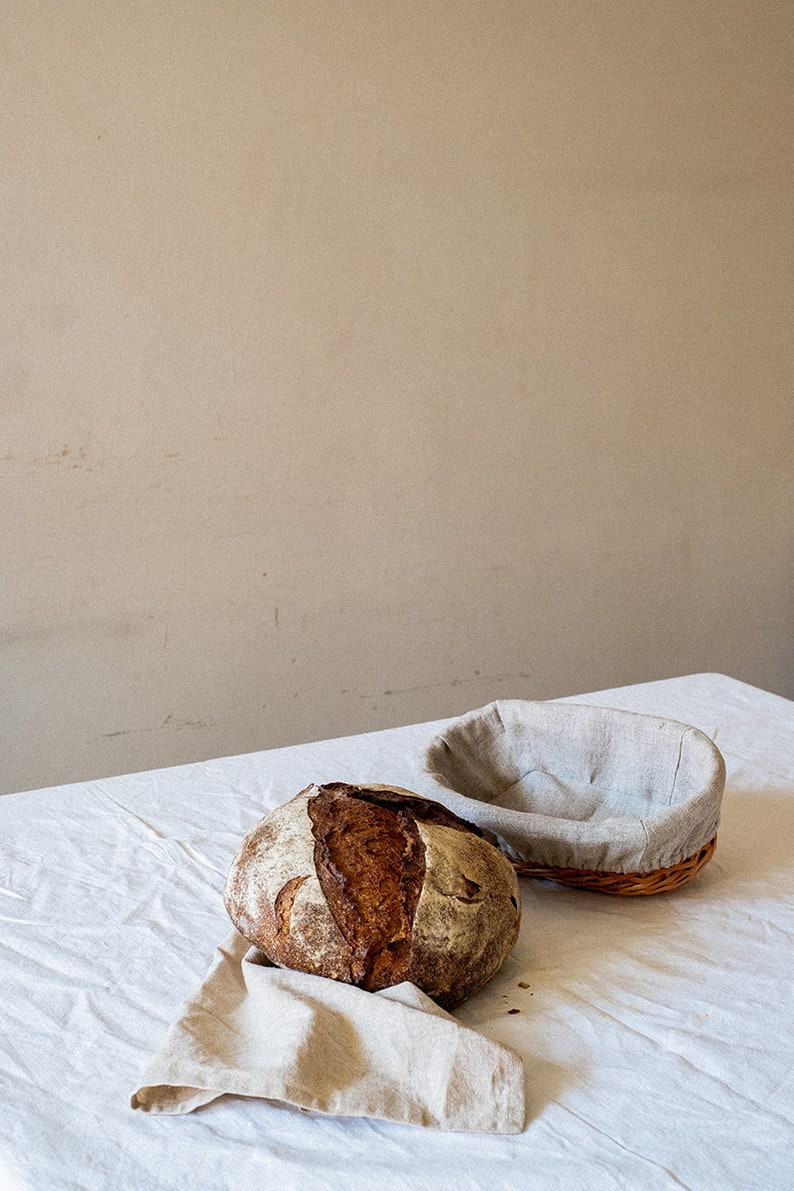 May include: A freshly baked loaf of sourdough bread sits on a white linen tablecloth, next to a brown wicker bread basket lined with a light gray linen cloth.