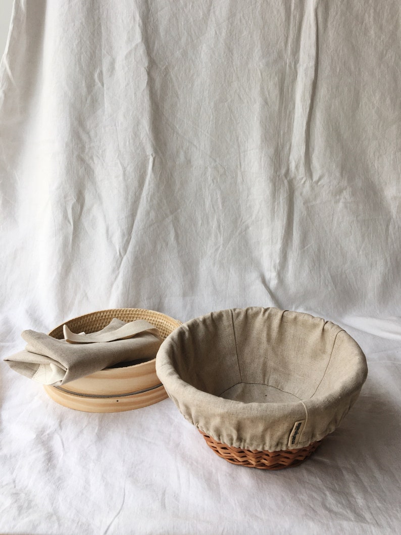 May include: A set of two beige linen-lined bread baskets. One basket is round and woven, the other is a wooden round with a removable linen liner.