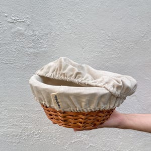 May include: A round, woven brown basket with a beige linen liner and cover. The basket is held by a hand. The label on the basket reads "mariamelia".