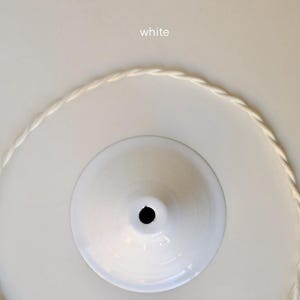 May include: A white ceiling canopy with a central circular opening, accompanied by a matching white twisted fabric electrical cord arranged in a circular shape. The word white is printed in small text above the canopy component.
