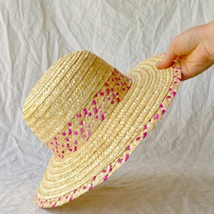 Puede incluir: Un sombrero de paja de ala ancha con un ribete tejido rosa. El sombrero est&aacute; hecho de paja natural y tiene una ala ligeramente ca&iacute;da.