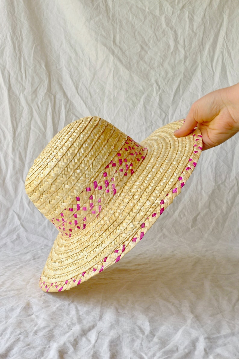 Puede incluir: Un sombrero de paja de ala ancha con un ribete tejido rosa. El sombrero est&aacute; hecho de paja natural y tiene una ala ligeramente ca&iacute;da.