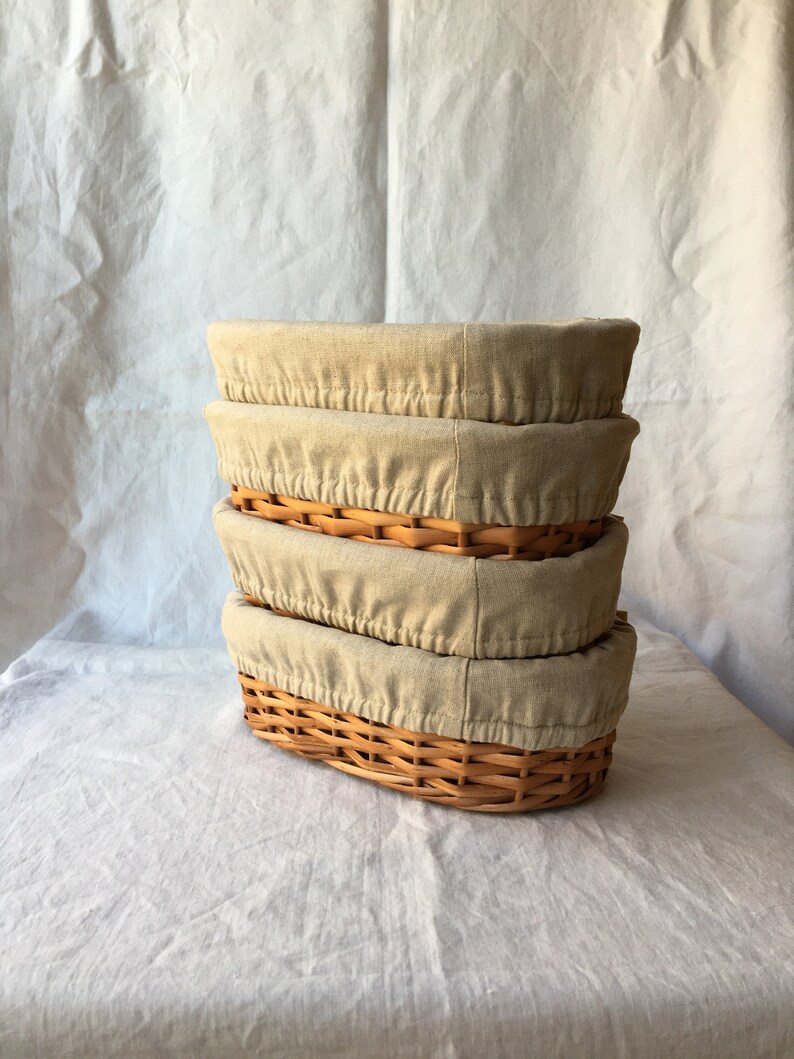 May include: Four oval wicker baskets with beige linen liners stacked on top of each other.