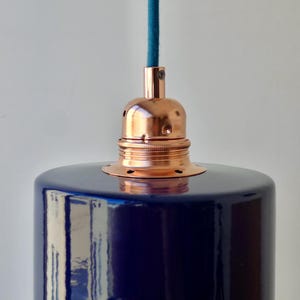 May include: A pendant light fixture featuring a glossy, deep navy blue cylindrical shade. The top of the shade is fitted with a polished copper metal socket holder, which connects to a textured teal blue fabric cord extending upward.