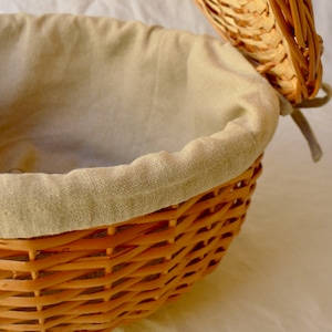 Pode incluir: Uma cesta de p&atilde;o redonda de vime com um forro de linho bege. A tampa est&aacute; aberta e a cesta est&aacute; vazia.