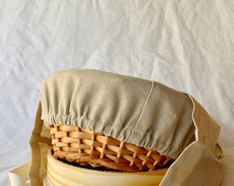 May include: A beige linen cover for a round wooden basket. The cover has a drawstring closure and is made of a natural fabric. The basket is made of woven wood and has a natural finish.