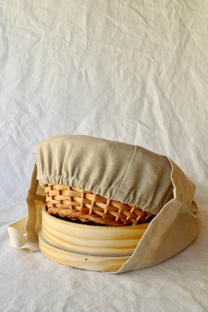 May include: A beige linen cover for a round wooden basket. The cover has a drawstring closure and is made of a natural fabric. The basket is made of woven wood and has a natural finish.