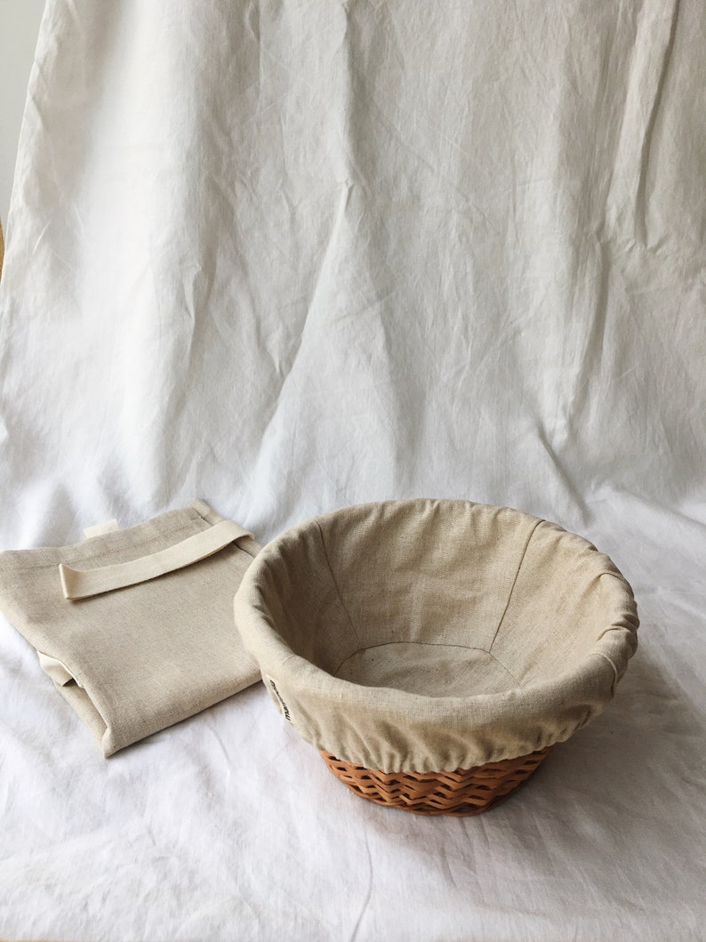 May include: A beige linen-lined wicker bread basket with a matching beige linen cloth on a white surface.
