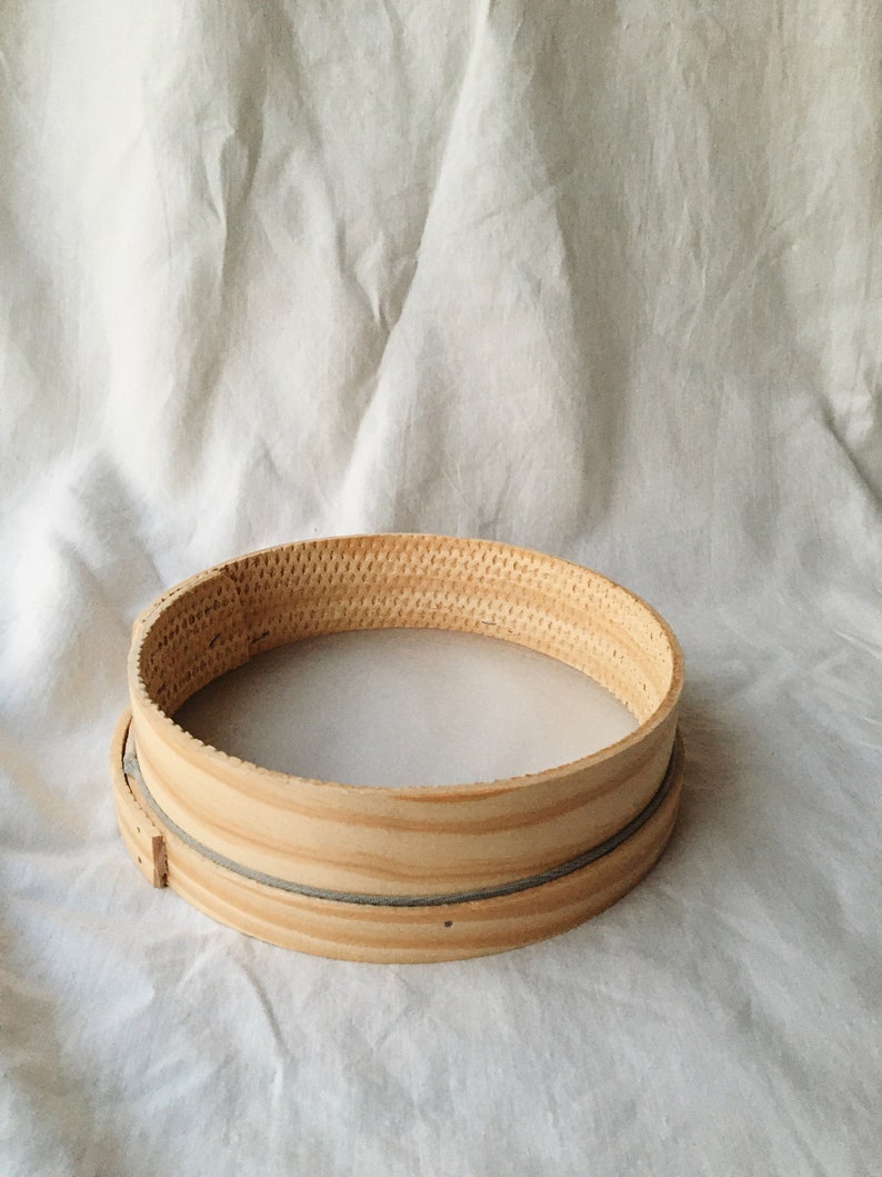 May include: A round wooden sieve with a woven mesh bottom. The sieve is made of light brown wood and has a natural finish.