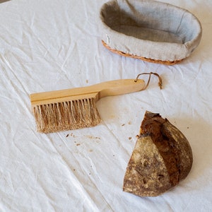 May include: A wooden handled brush with natural bristles, a loaf of bread, and a wicker bread basket with a linen liner on a white tablecloth.