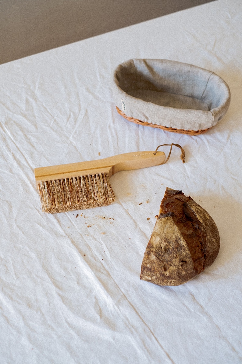 May include: A wooden handled brush with natural bristles, a loaf of bread, and a wicker bread basket with a linen liner on a white tablecloth.